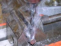 Twist bit drilling into a steel rod, wreathed in smoke from burning lubricant.
